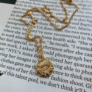 May include: Gold necklace with a round pendant featuring a stylised eye with a single diamond. The pendant is surrounded by a sunburst design with small stars.