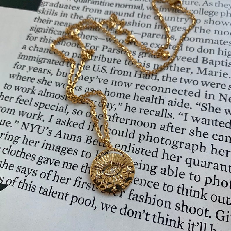 May include: Gold necklace with a round pendant featuring a stylised eye with a single diamond. The pendant is surrounded by a sunburst design with small stars.
