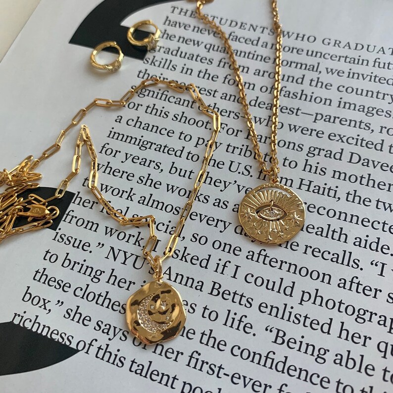 May include: Gold chain necklace with two pendants. One pendant is a round charm with a sun and an eye design. The other pendant is a crescent moon charm with a face and a star design.