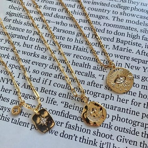 May include: Three gold necklaces with charms. The charms are a crescent moon with stars, a star with a sunburst, and an eye with stars. The necklaces are on a book with text about a fashion shoot.