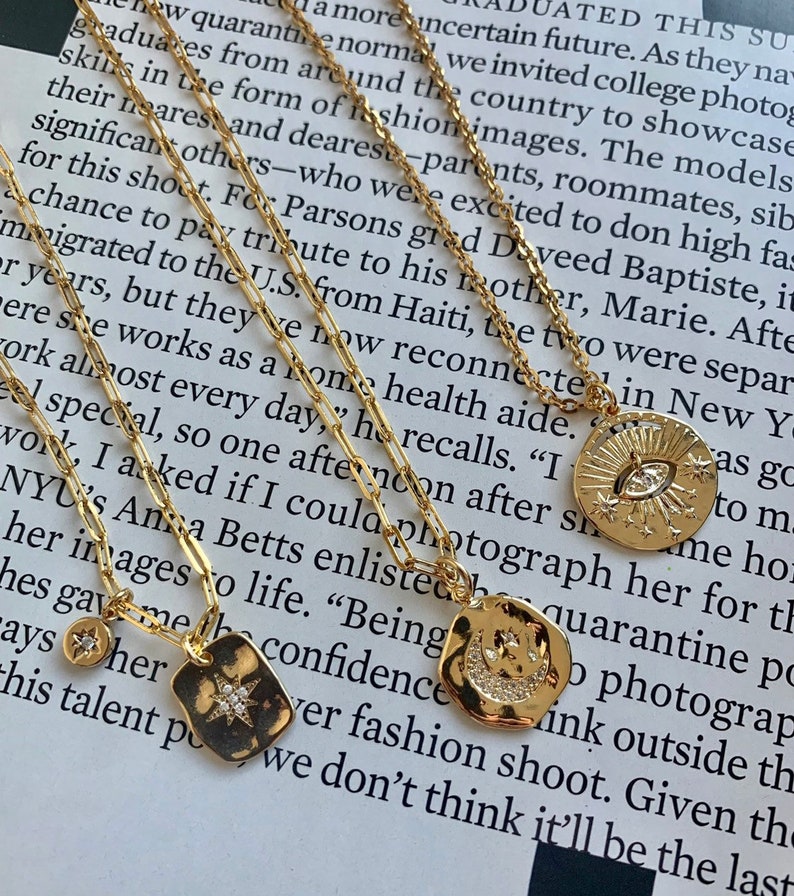 May include: Three gold necklaces with charms. The charms are a crescent moon with stars, a star with a sunburst, and an eye with stars. The necklaces are on a book with text about a fashion shoot.