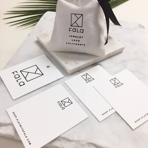 May include: A white fabric bag with a black ribbon closure. The bag has a black logo with the text "Fala Jewelry Love California". There are four white business cards with a black logo that says "Fala" and the text "Shop PlayWithFala.com".