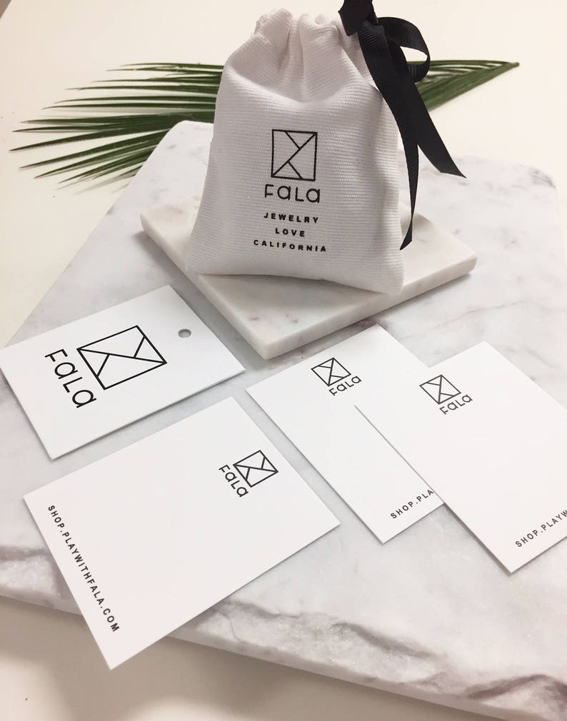 May include: A white fabric bag with a black ribbon closure. The bag has a black logo with the text "Fala Jewelry Love California". There are four white business cards with a black logo that says "Fala" and the text "Shop PlayWithFala.com".