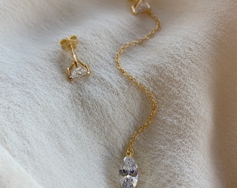Puede incluir: Un par de pendientes de oro con un diamante colgante de forma marquesa y un diamante triangular más pequeño. Los pendientes están sobre un fondo de tela blanca.
