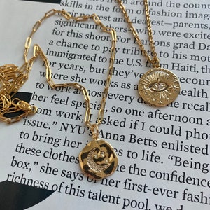 May include: A gold chain necklace with two pendants. One pendant is a round, hammered gold disc with a crescent moon and stars. The other pendant is a round, hammered gold disc with an eye and stars. The necklace is on a white surface with text in the background.