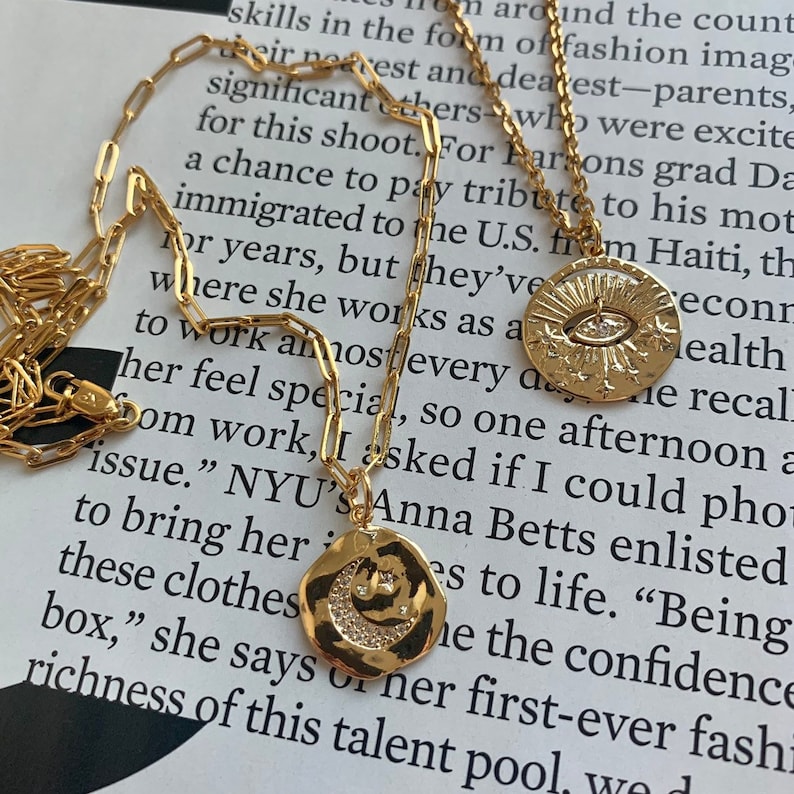 May include: A gold chain necklace with two pendants. One pendant is a round, hammered gold disc with a crescent moon and stars. The other pendant is a round, hammered gold disc with an eye and stars. The necklace is on a white surface with text in the background.