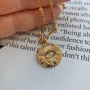 May include: Gold necklace with a round pendant featuring an eye design with small sparkling stones. The pendant has a textured surface and a sunburst design around the eye.