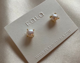 May include: A pair of pearl earrings with gold settings on a white card with the text "Faïla" and "www.playwithfaïla.com".