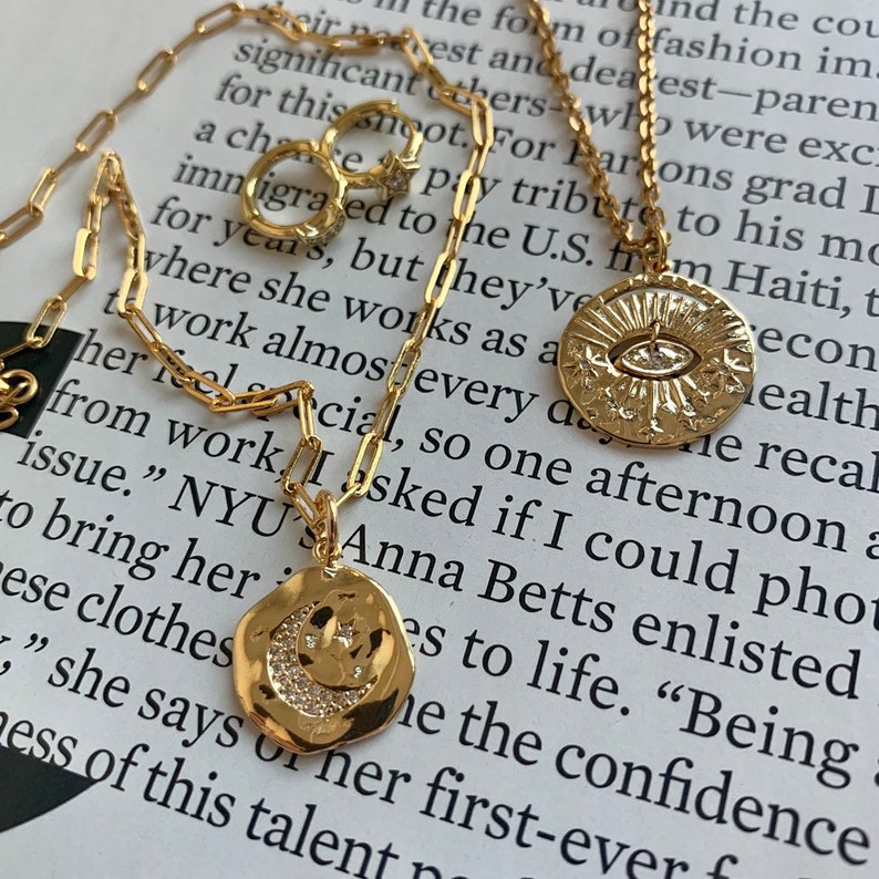 May include: A gold chain necklace with two pendants. One pendant is a crescent moon with a star and crystals. The other pendant is a sun with an eye and crystals. The necklace is on a book with text about fashion and design.