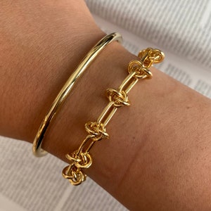 May include: Two gold bracelets on a person's wrist. One bracelet is a simple, thin bangle. The other bracelet is a thick chain with linked circles.