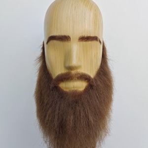 Op de afbeelding: Een houten hoofd met een volle bruine baard en snor. Het hoofd is gemaakt van lichtgekleurd hout, met bruine wenkbrauwen en een gebeeldhouwd gezicht. De baard is lang en vol, met een natuurlijk, getextureerd uiterlijk. Het hoofd is op een zwarte standaard gemonteerd.