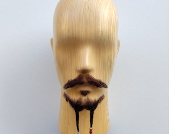 Può includere: Una testa di manichino in legno con barba e baffi marrone scuro. La barba ha due trecce, ognuna decorata con piccole perline verdi, rosse e dorate. La testa è montata su un treppiede nero, su sfondo bianco.