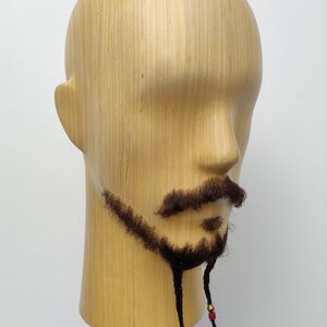 May include: A wooden mannequin head with a brown braided beard and mustache. The beard is tied with colorful beads.