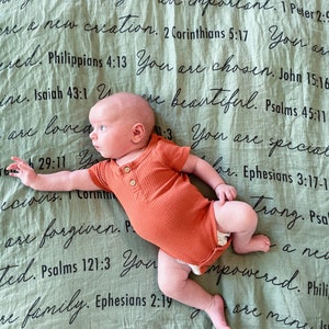 May include: A baby wearing a burnt orange onesie lies on a green blanket with black text. The text includes bible verses and phrases like "You are loved", "You are special", and "You are unique".