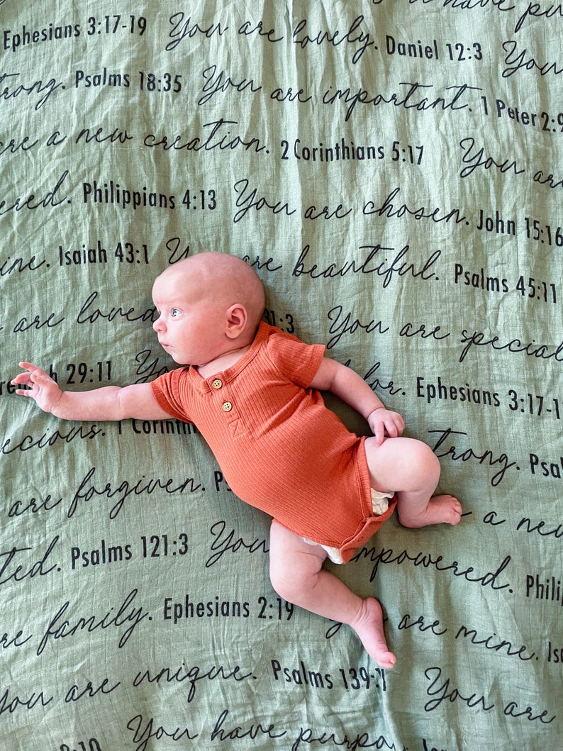 May include: A baby wearing a burnt orange onesie lies on a green blanket with black text. The text includes bible verses and phrases like "You are loved", "You are special", and "You are unique".