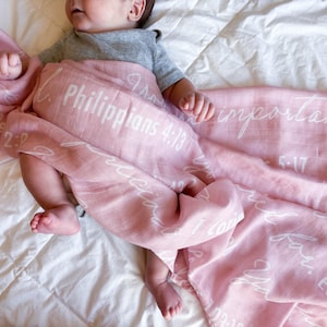 May include: A pink muslin swaddle blanket with white lettering. The blanket features various bible verses, including Philippians 4:13 and 1 Corinthians 13:4-7.