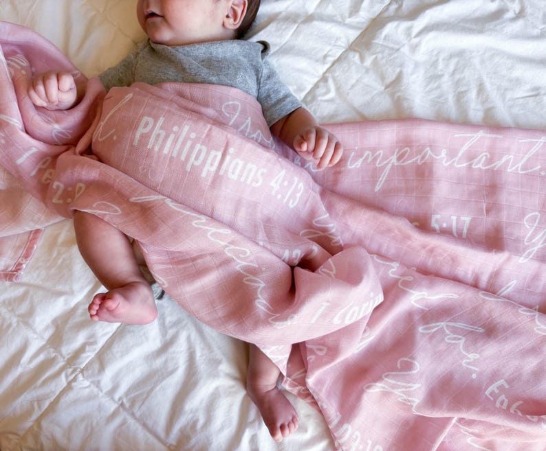 May include: A pink muslin swaddle blanket with white lettering. The blanket features various bible verses, including Philippians 4:13 and 1 Corinthians 13:4-7.