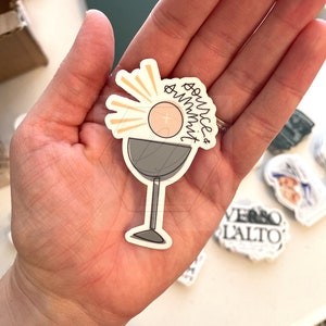 May include: A die-cut sticker featuring a gray chalice with a pale peach-colored host above it, surrounded by light orange radiating lines. The text "source & summit" is written in black cursive script to the right of the host and chalice.