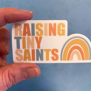 May include: A white sticker with the text "Raising Tiny Saints" in colorful block letters. A rainbow with orange, yellow, and blue stripes is on the right side of the sticker.