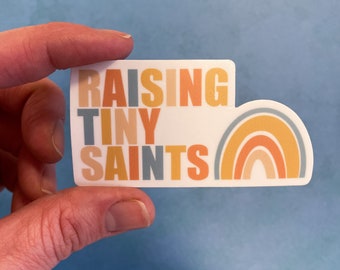May include: A rectangular sticker with a rounded right side featuring the text RAISING TINY SAINTS in a stacked, multi-colored sans-serif font. To the right of the text is a simple rainbow graphic composed of four arched bands in muted tones.