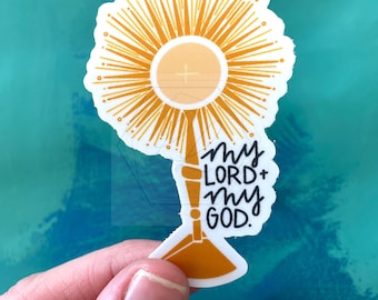 May include: A die-cut sticker featuring a gold-colored monstrance with a central host marked by a cross, surrounded by radiating sunburst lines. The text "my LORD + my GOD." is written in black, hand-lettered script on the right side of the base.