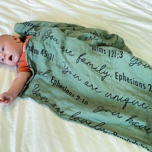 May include: A green muslin swaddle blanket with black text that reads "You are family. Ephesians 2:1" and "You are unique. Psalms 121:3". A baby is lying on a white sheet, partially covered by the blanket.