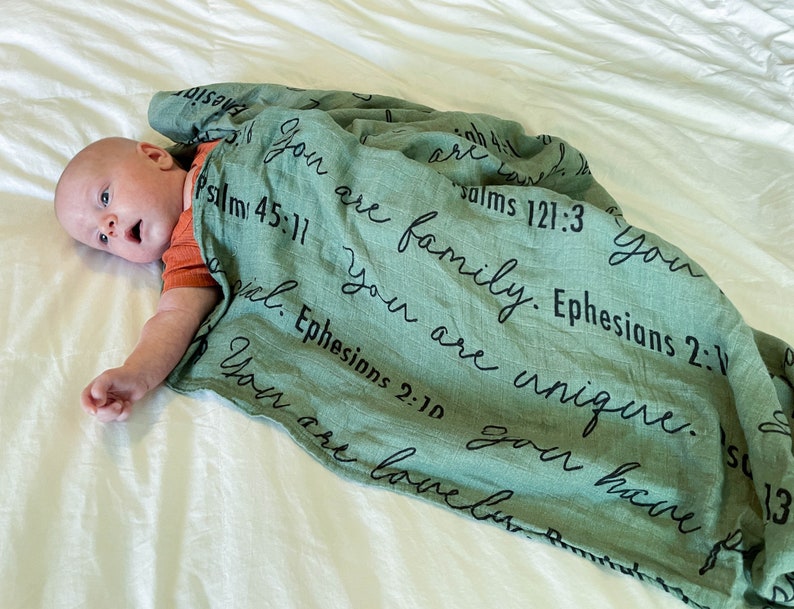 May include: A green muslin swaddle blanket with black text that reads "You are family. Ephesians 2:1" and "You are unique. Psalms 121:3". A baby is lying on a white sheet, partially covered by the blanket.