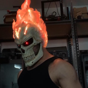 Op de afbeelding: Een schedelmasker met een gebeeldhouwd vlammenstuk dat oplicht met een intern oranje licht. Het masker heeft diepe, donkere oogkassen die worden verlicht door heldere rode LED-lampjes, een realistische botkleurige afwerking en een kaak.