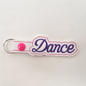 Puede incluir: Un llavero blanco con la palabra Dance bordada en letras cursivas de color morado. El llavero cuenta con un botón a presión rosa, costuras rosas a lo largo de los bordes y una anilla de metal plateado en un extremo para sujetar llaves.