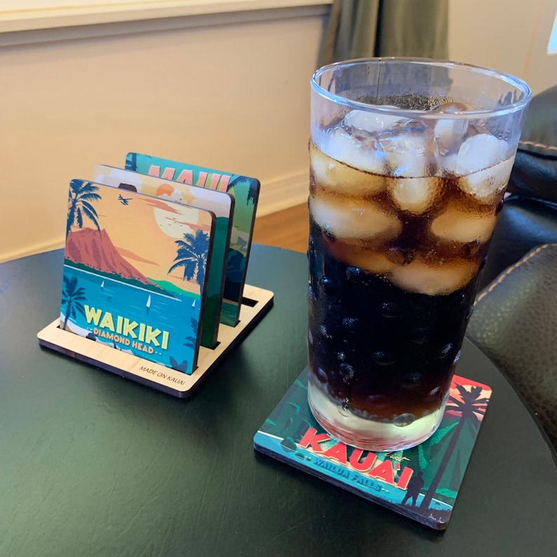 May include: A set of square wooden coasters featuring colorful illustrations of Hawaiian landmarks including Waikiki Diamond Head and Kauai Wailua Falls, stored in a wooden holder, with a tall glass of dark iced beverage resting on one.