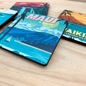 May include: A square coaster featuring a graphic illustration of a tropical landscape with blue mountains, a sailboat, and palm trees. The text MAUI The Valley Isle is printed in pink and white. Other coasters with similar travel-themed designs are visible.