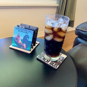 May include: A black table with a glass of cola with ice on a coaster that says "Beach Bus". There is a wooden coaster holder with four coasters on it. The coasters have images of a rooster, a chicken, and the words "Welcome to Kauai" and "The Chicken Patrol".