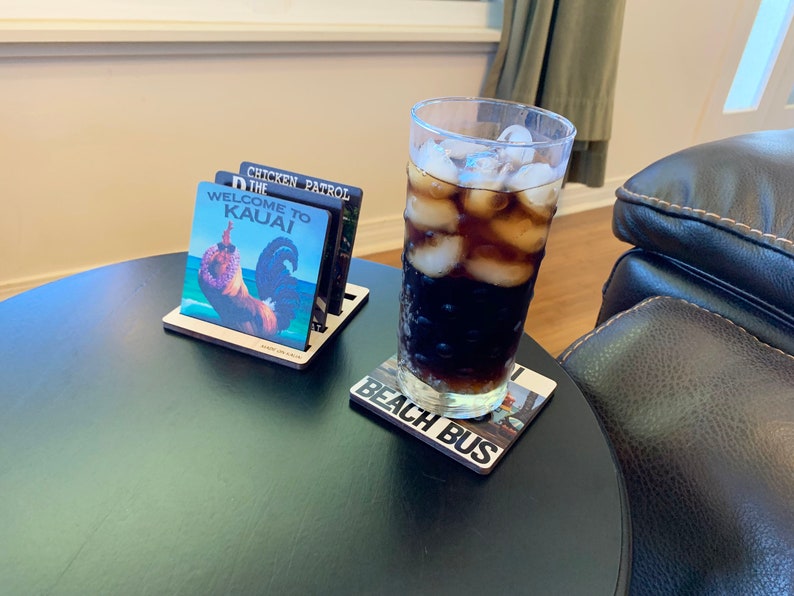 May include: A black table with a glass of cola with ice on a coaster that says "Beach Bus". There is a wooden coaster holder with four coasters on it. The coasters have images of a rooster, a chicken, and the words "Welcome to Kauai" and "The Chicken Patrol".