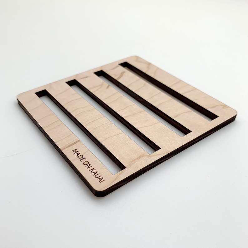 May include: A square light-colored wooden trivet featuring four parallel rectangular cutouts. The surface displays a natural wood grain pattern and the text MADE ON KAUAI is laser-engraved into the bottom left corner of the wooden frame.