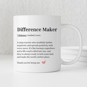 May include: White ceramic mug with a black and white design. The mug features the definition of "Difference Maker" as a ninja warrior who stealthily battles negativity and spreads positivity. The text includes "Thank you for being one" with a small pink heart.
