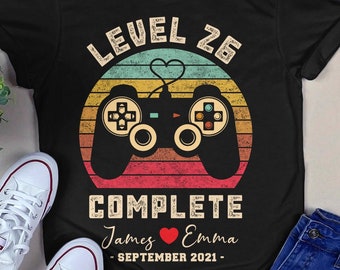 Op de afbeelding: Zwart T-shirt met een retro-stijl videogamecontroller afbeelding. De tekst "Level 25 Complete" staat boven de controller, en de tekst "James ♥ Emma - SEPTEMBER 2021 -" staat onder de controller.