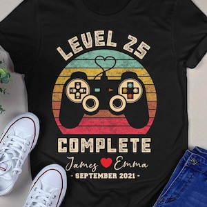 May include: Black t-shirt with a retro-style video game controller graphic. The text "Level 25 Complete" is above the controller, and the text "James ♥ Emma - September 2021 -" is below the controller.