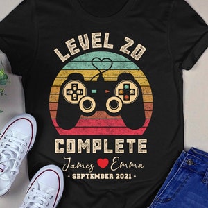 May include: Black t-shirt featuring a retro-style video game controller graphic. The text "Level 20 Complete" is above the controller, and the text "James ♥ Emma - September 2021 -" is below the controller.