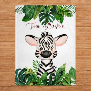 May include: White blanket featuring a watercolor illustration of a zebra with black and white stripes, surrounded by green tropical leaves. The name "Tom Haskin" is written in burgundy script across the top.