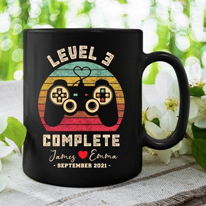 May include: Black coffee mug with a retro-style design featuring a video game controller and the text "Level 3 Complete" with the names "James & Emma" and the date "September 2021".