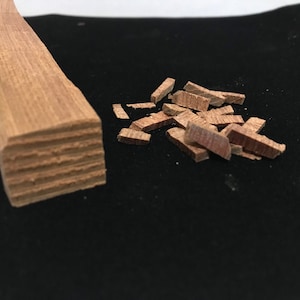 Könnte beinhalten: Nahaufnahme einer Holzleiste mit quadratischem Ende. Die Leiste besteht aus mehreren Holzschichten und wurde in kleine Stücke geschnitten. Die Stücke liegen auf einer schwarzen Oberfläche verstreut.
