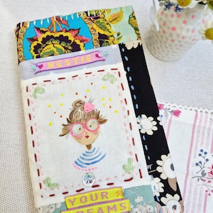 Könnte beinhalten: Eine rechteckige Stofftasche mit Patchwork-Design aus Blumen- und Schwarz-Weiß-Mustern. Das mittlere Feld zeigt eine gestickte Illustration einer Person mit rosa Brille und Dutt sowie den Text "BESTIE" und "YOUR DREAMS".