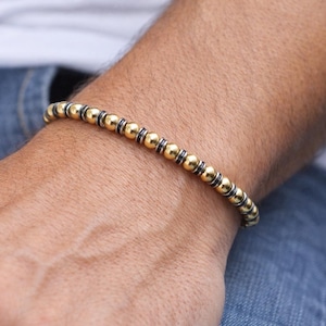 May include: Gold beaded bracelet with black accents. The bracelet is made of small, round gold beads separated by black spacers. The bracelet is worn on a wrist, with a blue jeans and white shirt in the background.