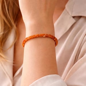 May include: A close-up of a wrist wearing a square-beaded amber bracelet. The bracelet is a warm, orange-brown color and appears to be made of small, evenly sized beads. The background is a soft, neutral color, with a hint of a collared shirt.