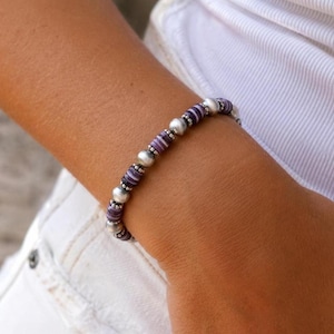 May include: A beaded bracelet featuring alternating purple and silver beads, interspersed with round, white pearls. The bracelet is worn on a wrist, with a white shirt visible in the background. The jewelry is a mix of colors and textures.
