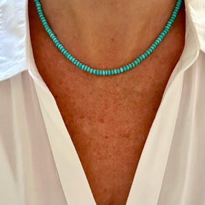 May include: A turquoise beaded necklace resting on a person's neck. The necklace is composed of small, round, turquoise beads. The person is wearing a white collared shirt. The necklace is a simple, elegant piece of jewelry.