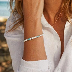 May include: A beaded bracelet featuring a repeating pattern of small, flat, circular white and turquoise beads. The bracelet is constructed with a series of white discs interspersed with occasional bright turquoise accents, creating a simple, uniform, and linear design around the wrist.