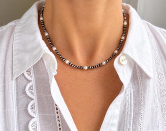 May include: A beaded necklace featuring alternating small dark metallic gray beads and larger round white pearls. The beads are arranged in a repeating pattern, with silver-toned spacers placed between the pearls and the metallic gray beads.