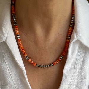 May include: A necklace made of orange and white beads with silver accents. The necklace is worn by a person with a white shirt.