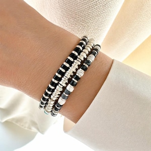 May include: Three beaded bracelets stacked on a wrist. The bracelets feature a mix of black, silver, and white beads. The beads are round and strung together. The background is a light-colored fabric.